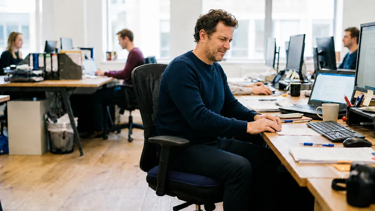 Homme assis confortablement à son bureau sur le coussin KinéConfort, les épaules relâchées et l’appui réparti